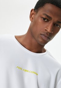Hvid kortærmet T-shirt i et struktureret stof med "FEEL CHALLENGED" i fed gul skrift. Rund halsudskæring, afslappet pasform.