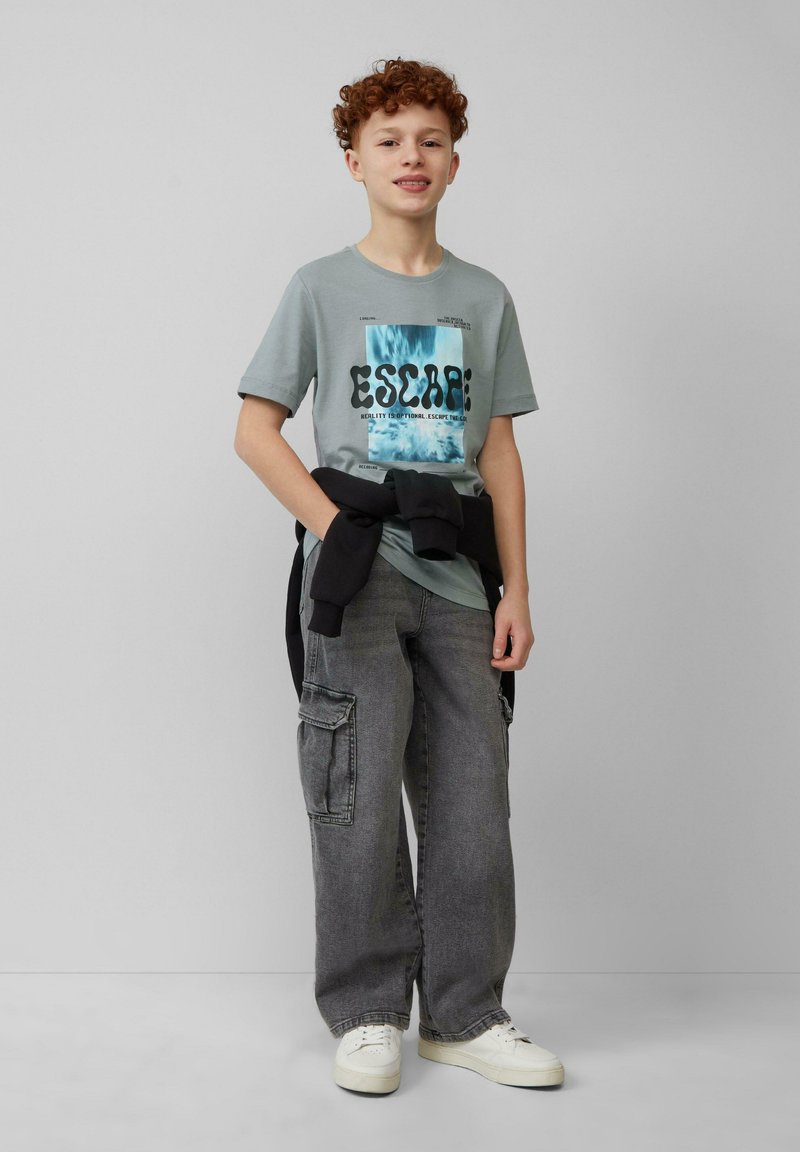 Grijze t-shirt met een blauw grafisch ontwerp en zwarte tekst, gecombineerd met losse grijze cargo-broeken en witte sneakers. Zwarte sweater gestrikt om de taille.