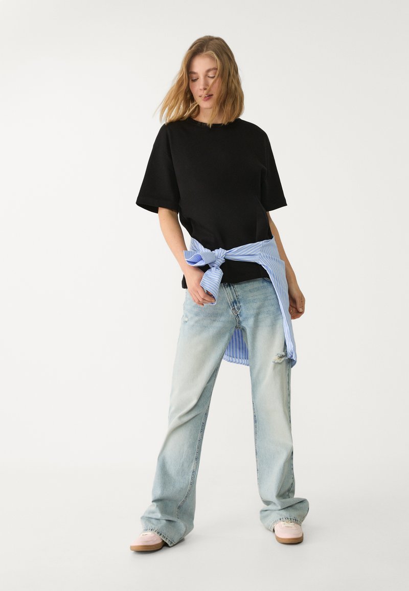 Sort oversized t-shirt parret med lyseblå, slidte wide-leg jeans. En lyseblå stribet skjorte bundet om livet tilføjer kontrast.