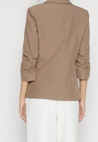 Brun strukturerad kavaj med en enkel bakslits, rullade ärmar, skräddarsydd passform och en mjuk yta, i kombination med vita vida byxor.