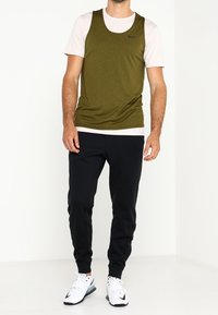 Débardeur de sport vert porté sur un t-shirt clair, associé à un jogging noir et des baskets blanches. Le débardeur arbore un petit logo noir.