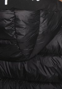 Svart pufferjacka med en kviltad textur, huva och vitt logo på toppen. Materialet ser glänsande och isolerat ut.