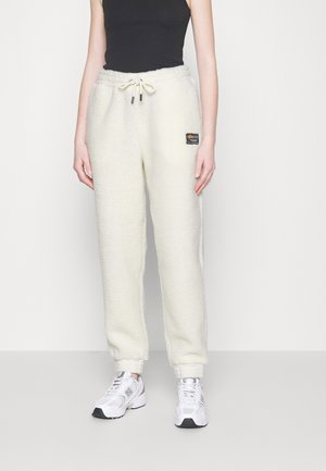 Cream-colored fleece joggers with an elastic waistband, drawstring closure, side pockets, and a logo patch on the left thigh.