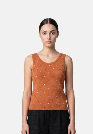 Femme portant un haut sans manches de couleur rouille avec un motif en losange et de petites décorations en perles, associé à un pantalon noir sur fond blanc.