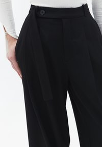 Zwarte op maat gemaakte broek met een hoge taille, voorzien van een zijstrik, enkele zwarte knoop en voorplooien voor extra structuur.