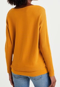 Mustard yellow ribbed sweater with long sleeves and a wide neckline, featuring a relaxed fit and side slits at the hem.