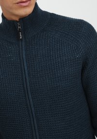 Námořnická svetr na zip s texturovaným pletením. Obsahuje vysoký límec a kontrastní zip. Hladká tkanina s mírně žebrovanou texturou.