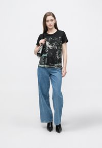 Čierne grafické tričko s motívom mačky, v kombinácii so svetlomodrými širokými džínsami a čiernymi členkovými topánkami na podpätku, doplnené čiernou taškou.