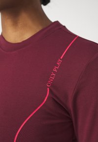 Burgundi sportinė marškinė su apvali iškirpte; priekyje yra ryškiai rožinė "ONLY PLAY" užrašas ir kreiva akcento linija.