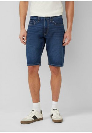 Man die donkerblauwe denimshorts draagt, witte sportsokken, witte sneakers met zwarte accenten en een wit T-shirt dat bij de taille is afgeknipt.