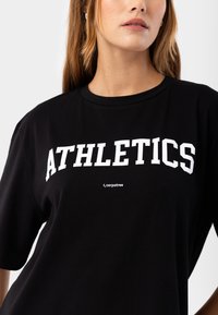Melna kokvilnas T-kreklu ar īsām piedurknēm, ar lielu baltu tekstu "ATHLETICS" krūšu daļā un mazu logotipu zem tā.