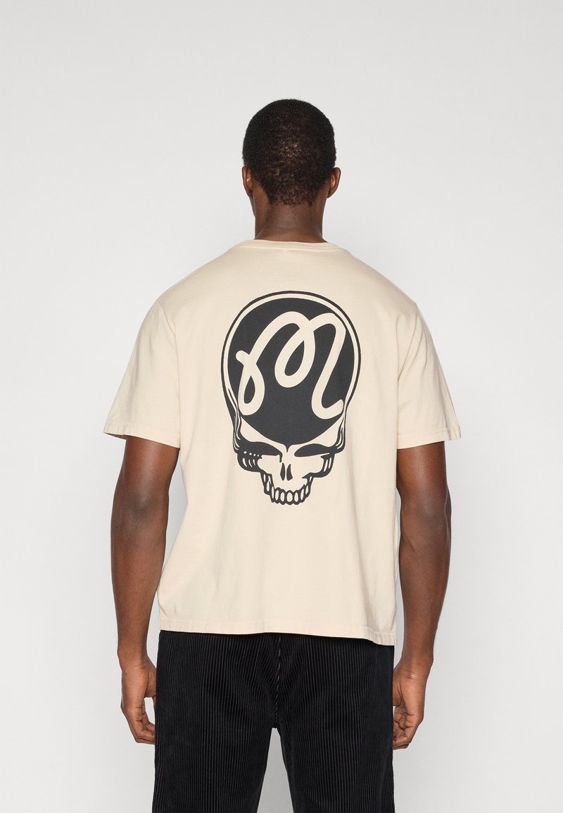 Μπεζ βαμβακερό μπλουζάκι με μεγάλο μαύρο σχέδιο κρανίου στην πλάτη. Κοντά μανίκια, λαιμόκοψη και άνετη εφαρμογή. Φοριούνται μαύρο παντελόνι κοτλέ.