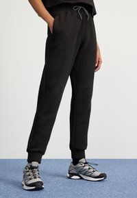 Pantalons de jogging noirs texturés avec une taille et des poignets côtelés, dotés de poches latérales et d'un cordon de serrage gris, associés à des chaussures de sport grises.