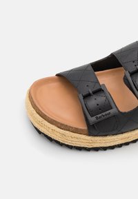 Sandale en cuir noir avec un design matelassé, deux boucles réglables, une semelle intérieure en synthétique marron, et une semelle en jute entourée de caoutchouc noir.