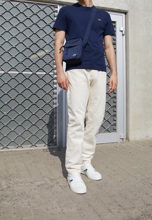 Mężczyzna ubrany w granatowy t-shirt, kremowe spodnie, białe trampki i granatową torbę na ramię, stojący na wybrukowanym podłożu obok metalowych drzwi z kratką.