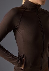 Chaqueta ajustada de color marrón, con cremallera y manga larga, con bolsillos laterales, fabricada en una tela suave y elástica. Detalles de costura visibles.