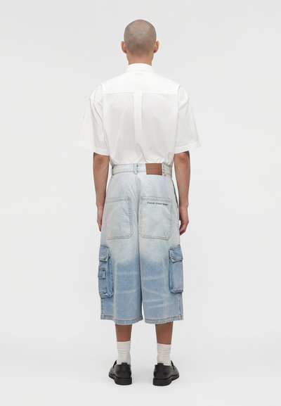 Άντρας με λευκό κοντομάνικο πουκάμισο, ανοιχτό μπλε φαρδιά τζιν σορτς με τσέπες cargo, λευκές κάλτσες και μαύρα παπούτσια, στέκεται με την πλάτη γυρισμένη.