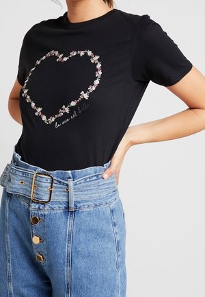Woman wearing black short-sleeve t-shirt with floral heart and "la vie est belle" text, paired with high-waisted blue jeans and a belt.