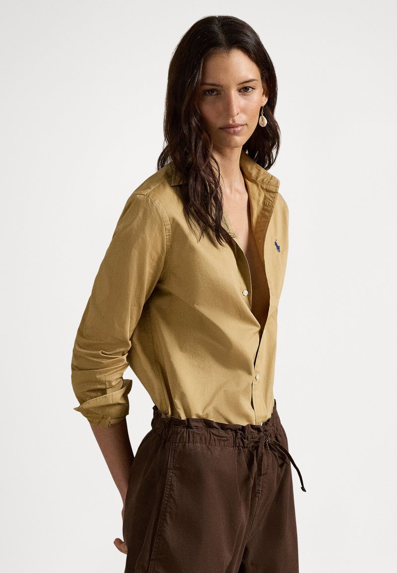 Žena s dlhými tmavými vlasmi, oblečená v hnedom košeľovom tričku a hnedých nohaviciach, stojaca s rukami za chrbtom na jednoduchom pozadí.