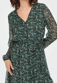 Donna che indossa un vestito floreale a maniche lunghe, blu petrolio e nero, con scollatura a V e cintura annodata in vita.