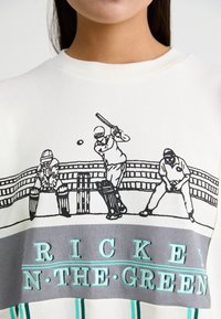 Λευκή μπλούζα με γραφικό σχέδιο κρίκετ σε μαύρο χρώμα με μπλε τόνους, απεικονίζοντας παίκτες σε δράση και την επιγραφή "ΚΡΙΚΕΤ ΣΤΟ ΠΡΑΣΙΝΟ."