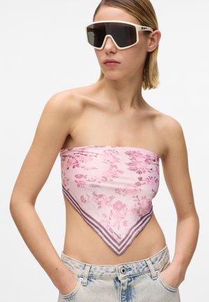 Γυναίκα που φοράει υπερμεγέθη γυαλιά ηλίου με λευκή σκελετό, ροζ μπαντάνα με λουλούδια και ανοιχτόχρωμο τζιν παντελόνι με τα χέρια στις τσέπες.