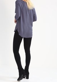 Loose-fitting, long-sleeve top in light gray, paired with black leggings and ankle boots. Soft fabric with a rounded hem and subtle drape.