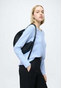 Czarna torebka o zaokrąglonym kształcie i fakturowanej powierzchni, noszona z jasnoniebieską koszulą z długim rękawem i czarnymi spodniami. Prosty design, minimalistyczne elementy metalowe.