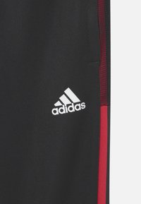 Černé sportovní kalhoty s červenými bočními akcenty, z texturované látky a s bílým logem Adidas na levé straně.