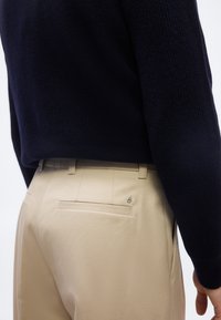 Personne portant un pantalon beige avec une poche arrière et un haut noir texturé à manches longues, vue de la taille vers le bas et de profil.