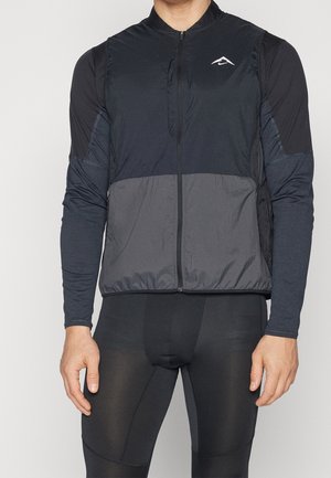 Mand iført en sort og mørkegrå ærmeløs vest med lynlås over en langærmet sort sportstrøje og sorte tætsiddende sportslangebukser.