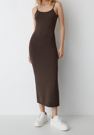 Robe maxi en satin marron avec des fines bretelles, silhouette ajustée et texture lisse, associée à des baskets plates blanches.