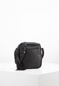 Svart lädercrossbody-väska med fyrkantig form, flätad rem, dragkedjestängning och en tofsdetalj. Slät textur och minimalistisk design.