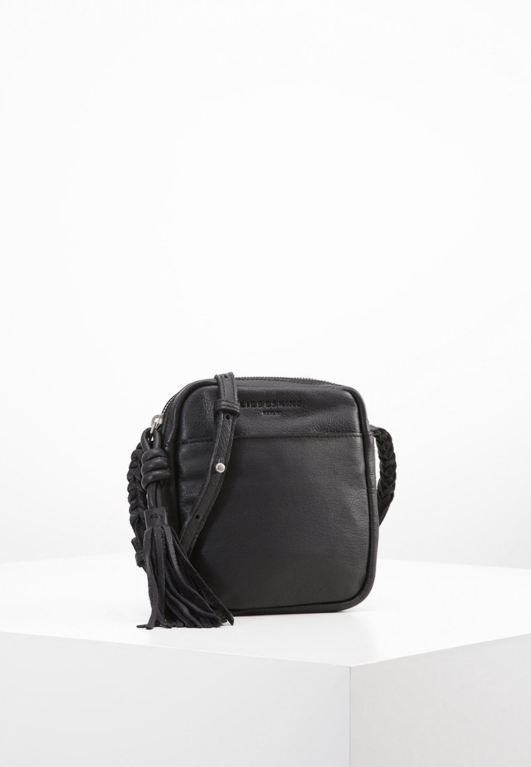 Svart lädercrossbody-väska med fyrkantig form, flätad rem, dragkedjestängning och en tofsdetalj. Slät textur och minimalistisk design.