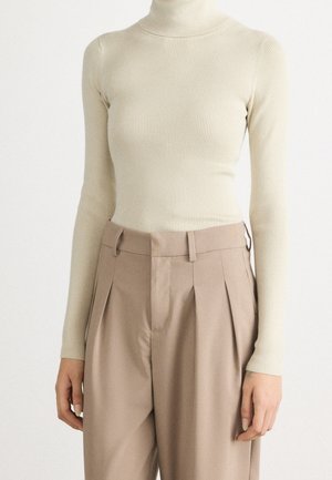 Personne portant un pull à col roulé crème ajusté et côtelé et un pantalon beige plissé taille haute, debout devant un fond uni.