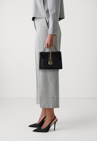 Czarna torebka z łańcuszkowym detalem, o uporządkowanym kształcie i złotych elementach, zestawiona z kraciastym garniturem i czarnymi szpilkami.