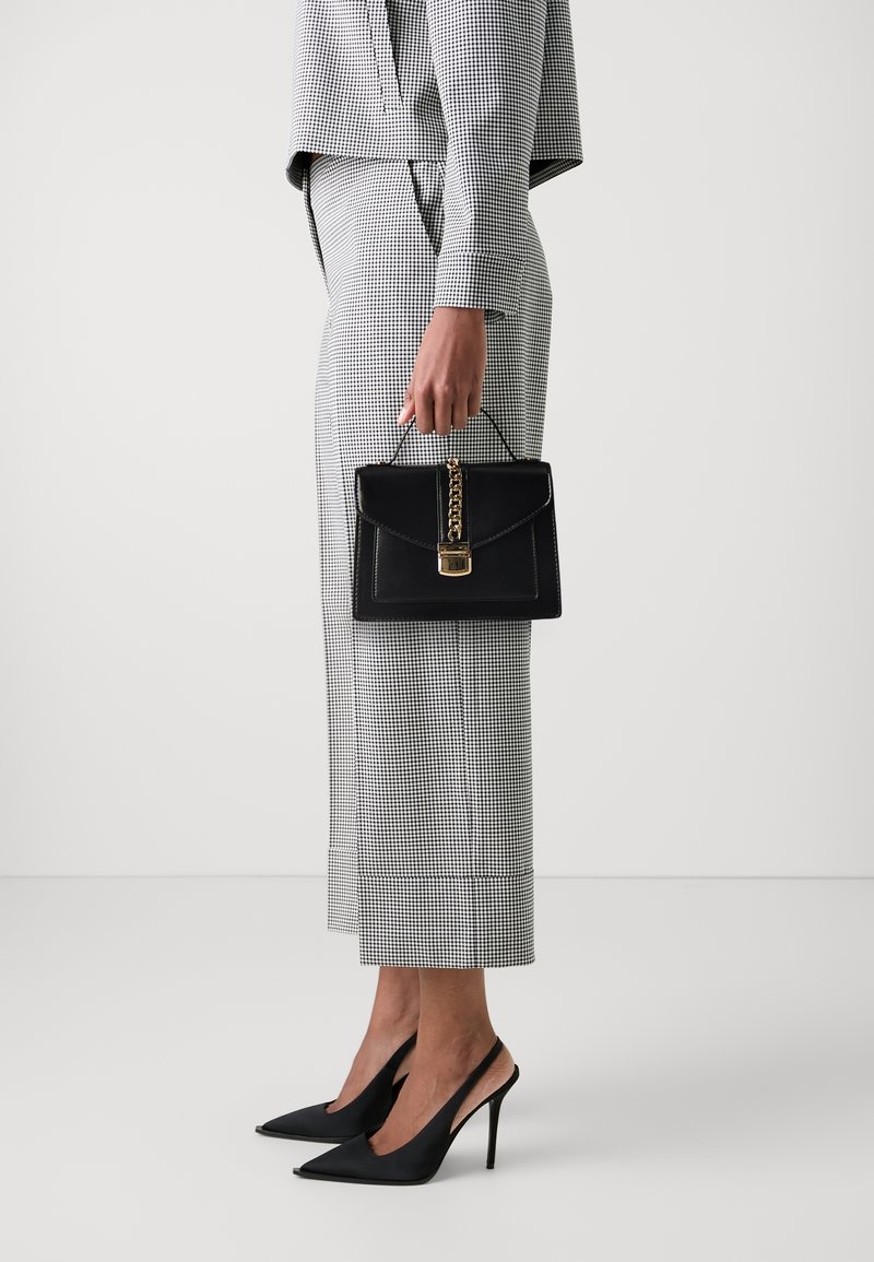 Czarna torebka z łańcuszkowym detalem, o uporządkowanym kształcie i złotych elementach, zestawiona z kraciastym garniturem i czarnymi szpilkami.
