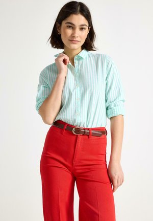 Donna con capelli corti e scuri che indossa una maglietta a righe verde chiaro e bianca e pantaloni rossi vivaci con una cintura marrone, in posa davanti a uno sfondo semplice.