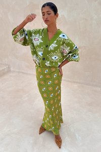 Cardigã verde de malha com padrões florais e de olhos, combinado com uma saia longa correspondente com motivos semelhantes. Sapatos castanhos de salto alto.