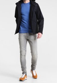 Marinblå huvtröja över en blå sweatshirts, ljusgrå jeans och orange/vita sneakers. Funktioner inkluderar dragkedja och sidofickor.