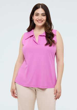 Mujer con cabello largo y oscuro sonriendo, vistiendo una blusa sin mangas rosa y pantalones beige sobre un fondo claro y liso.