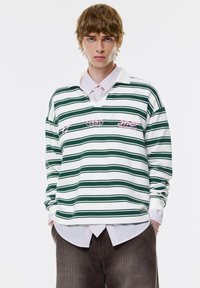 Biała i zielona w paski, długorękawowa koszulka polo z dekoltem w serek, z różowymi haftowanymi logo. Noszona na jasnoróżowej koszuli w paski.