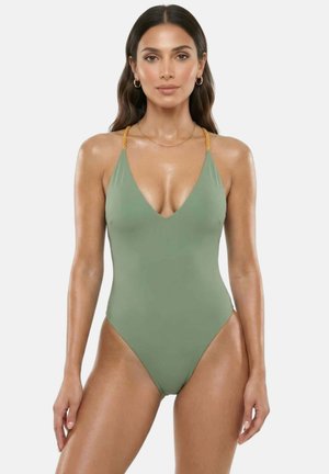 Frau mit langen dunklen Haaren, die einen grünen Einteiler mit gedrehten beigen Schulterriemen trägt und vor einem schlichten hellen Hintergrund steht.