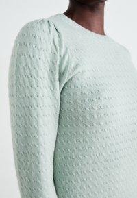 Pull en maille vert clair avec un motif texturé présentant des lignes diagonales subtiles et un détail froncé aux épaules. Col rond.