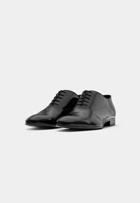 Un paio di scarpe oxford di pelle nera lucida con lacci chiusi, esposte su uno sfondo bianco semplice.