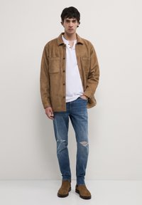 Giacca in suede marrone con due tasche frontali, indossata sopra un polo bianco. Jeans strappati azzurri e stivali in suede marrone completano l'outfit.