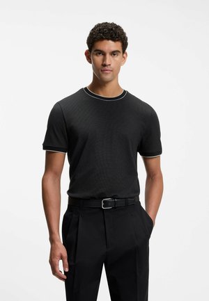 Mand med krøllet hår iført en sort t-shirt med korte ærmer og rund hals samt sorte bukser med bælte, står foran en ensfarvet hvid baggrund.