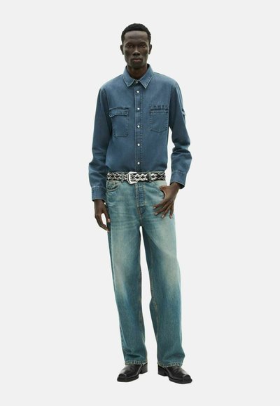 Camicia di denim blu scuro con bottoni sul davanti, due tasche sul petto; jeans azzurri a gamba larga con leggero sbiadimento e scarpe in pelle nera.