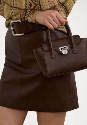 Brown leather handbag with a textured finish and silver hardware, held with a hand beside a brown leather skirt and patterned sweater.