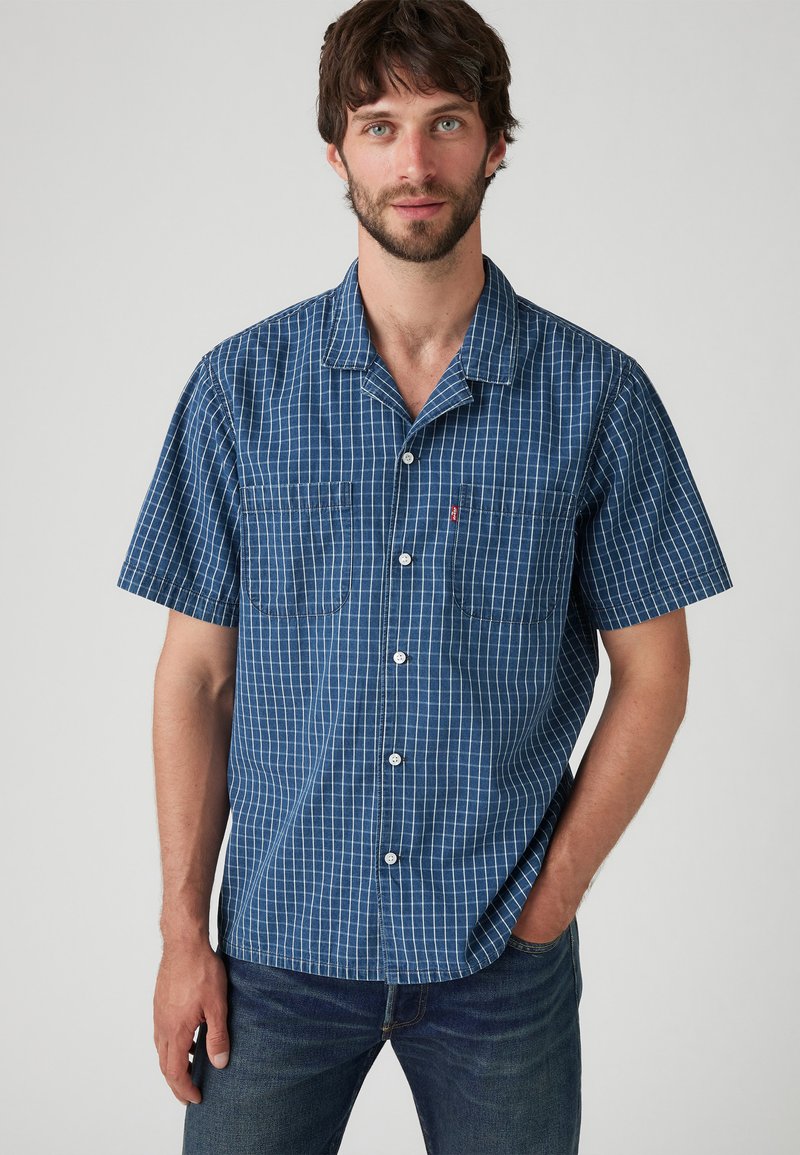 Camisa de cuadros azul de manga corta en una tela ligera, con cuello abotonado, dos bolsillos frontales y detalles de botones blancos.